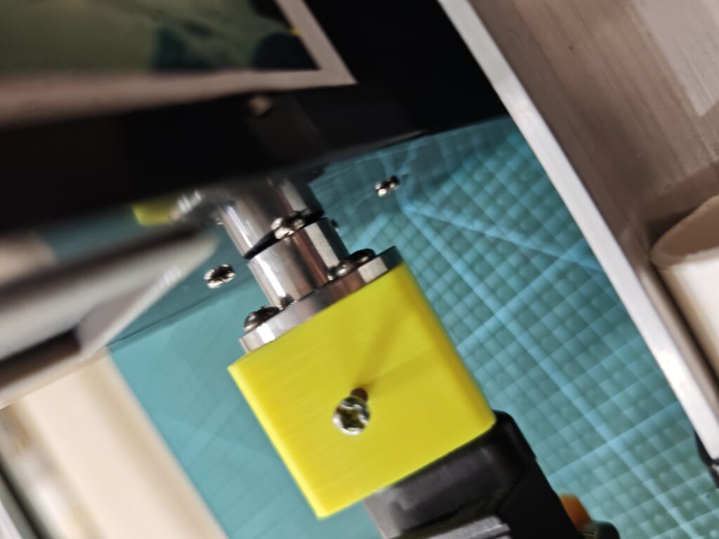 Close-up, angled view of a custom 3D-printed yellow adapter connecting a metal flanged coupler to the azimuth (AZ) motor shaft of an amateur radio antenna rotator, secured with 4 screws.